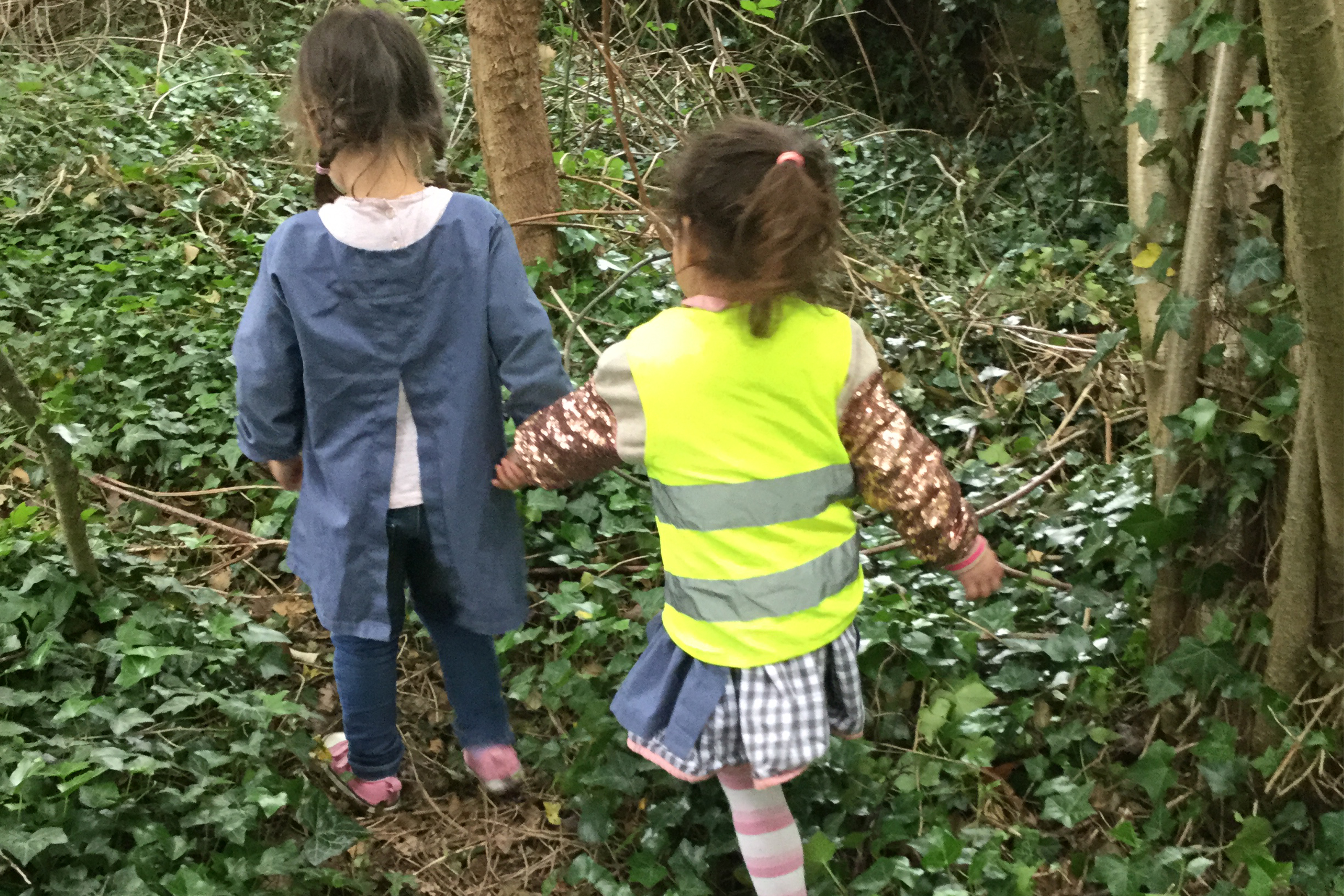 Two children walking hand in hand through the woods at Forest School